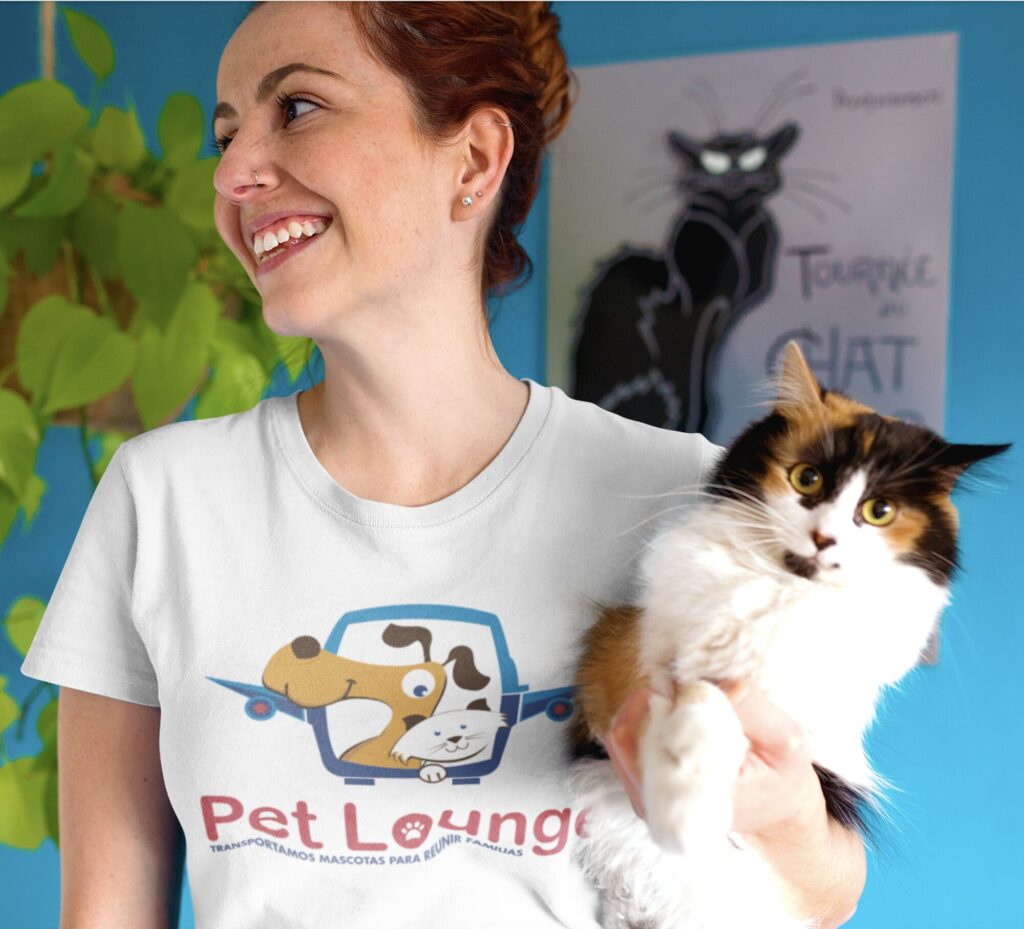 A smiling woman in a "Pet Lounge" T-shirt holding a fluffy calico cat in front of a blue wall with a cat poster.